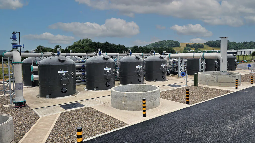 Numerous black cylindrical tanks interconnected by piping, arranged in rows within a fenced industrial area against a backdrop of rolling hills.