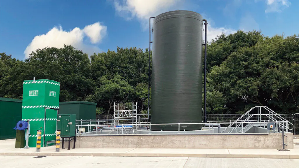 Industrial site with tanks, pipes, and green utility buildings; workers in high-visibility clothing are present.