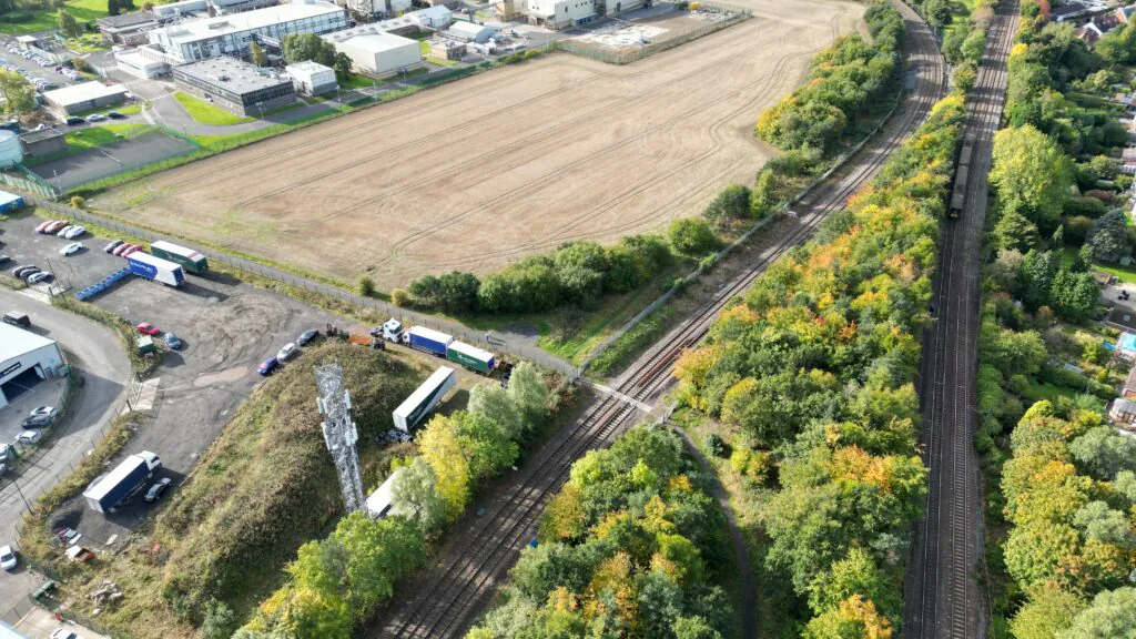 Aerial view of railway tracks running parallel to an open field with industrial buildings nearby, showing infrastructure development and surrounding greenery.