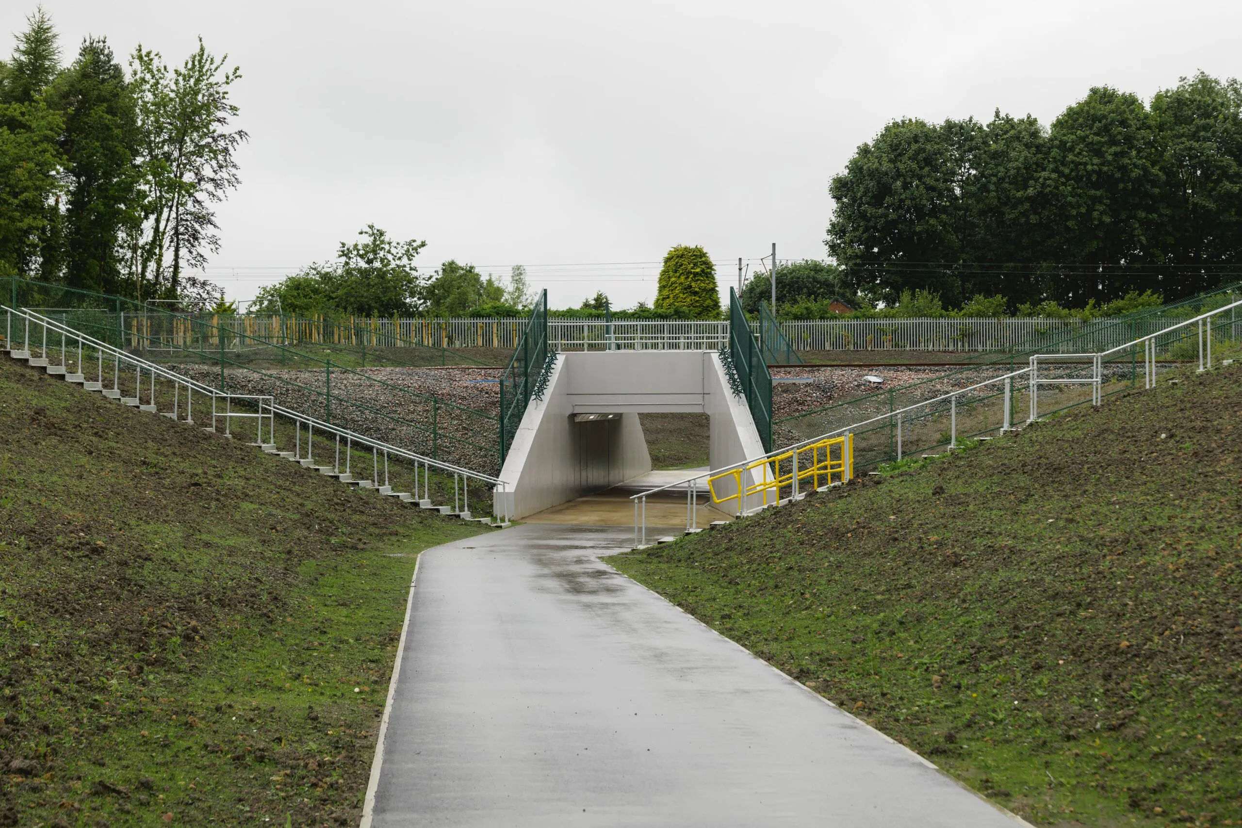 Newly constructed pedestrian underpass with railings on both sides leading to the tunnel entrance; wet pathway surrounded by grass and trees.