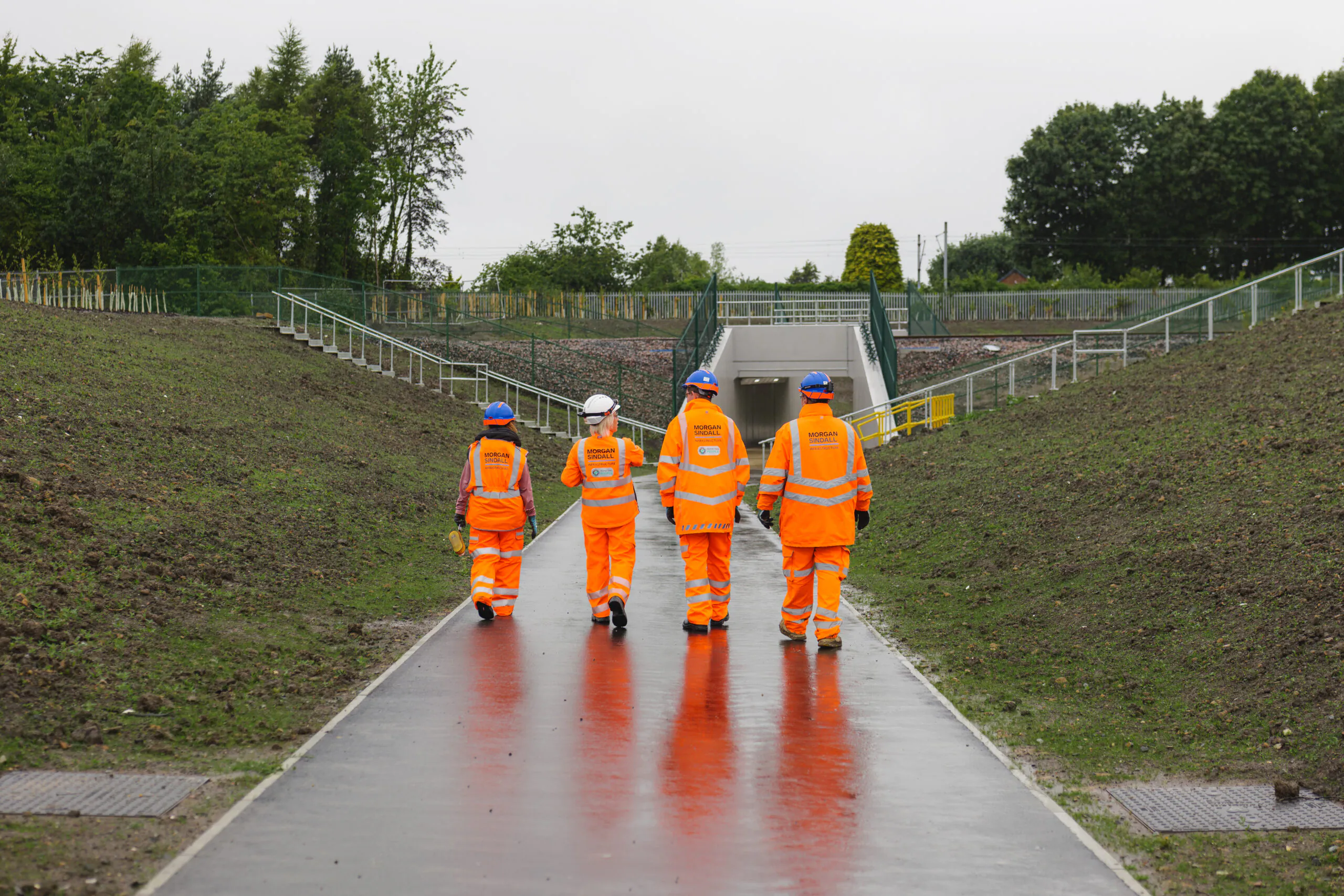 Four individuals in high-visibility clothing walking toward the pedestrian underpass on a rain-wet path reflecting their orange attire.