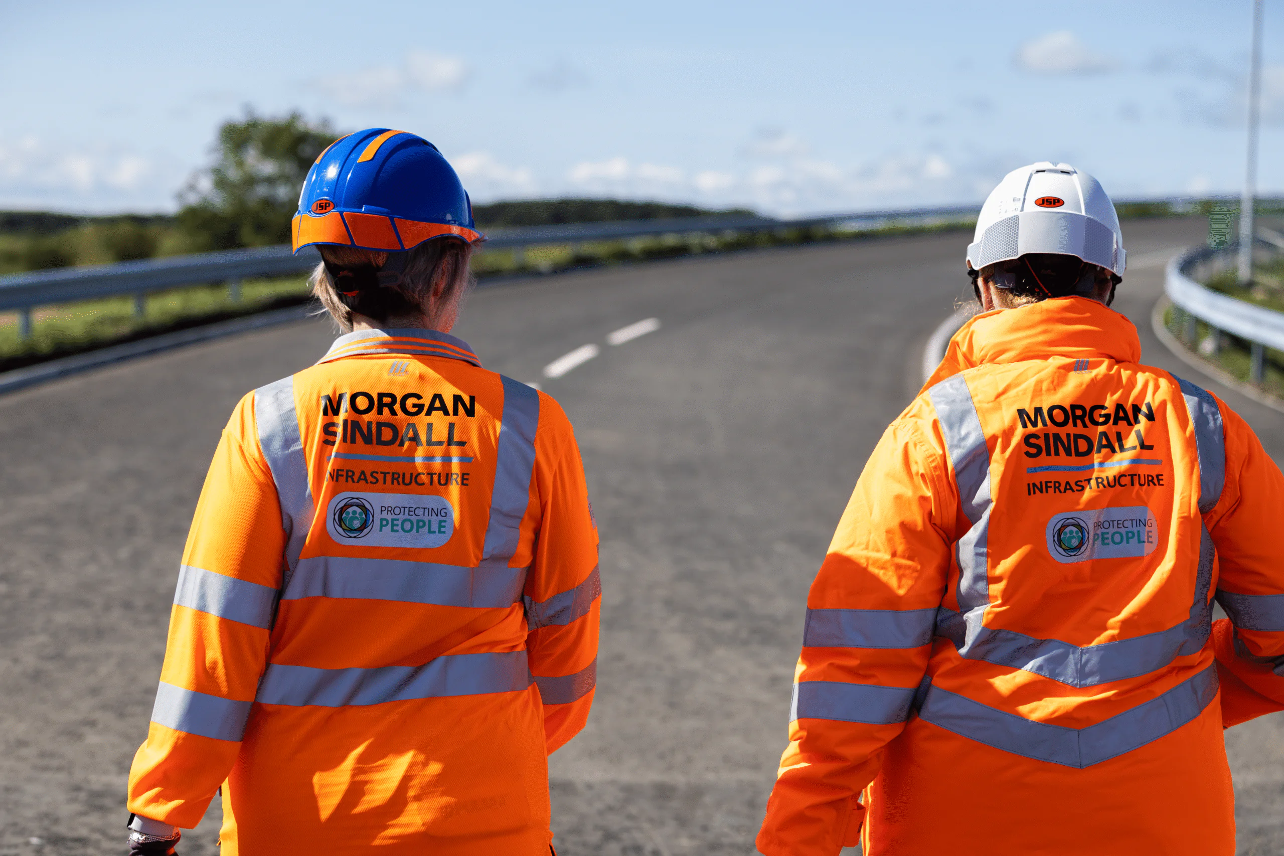 Two individuals walking down an empty road wearing orange high-visibility jackets labeled “Morgan Sindall Infrastructure.”