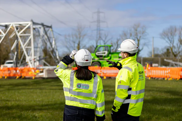 Two individuals in high-visibility jackets and white hard hats standing on a grassy construction site, one pointing into the distance.