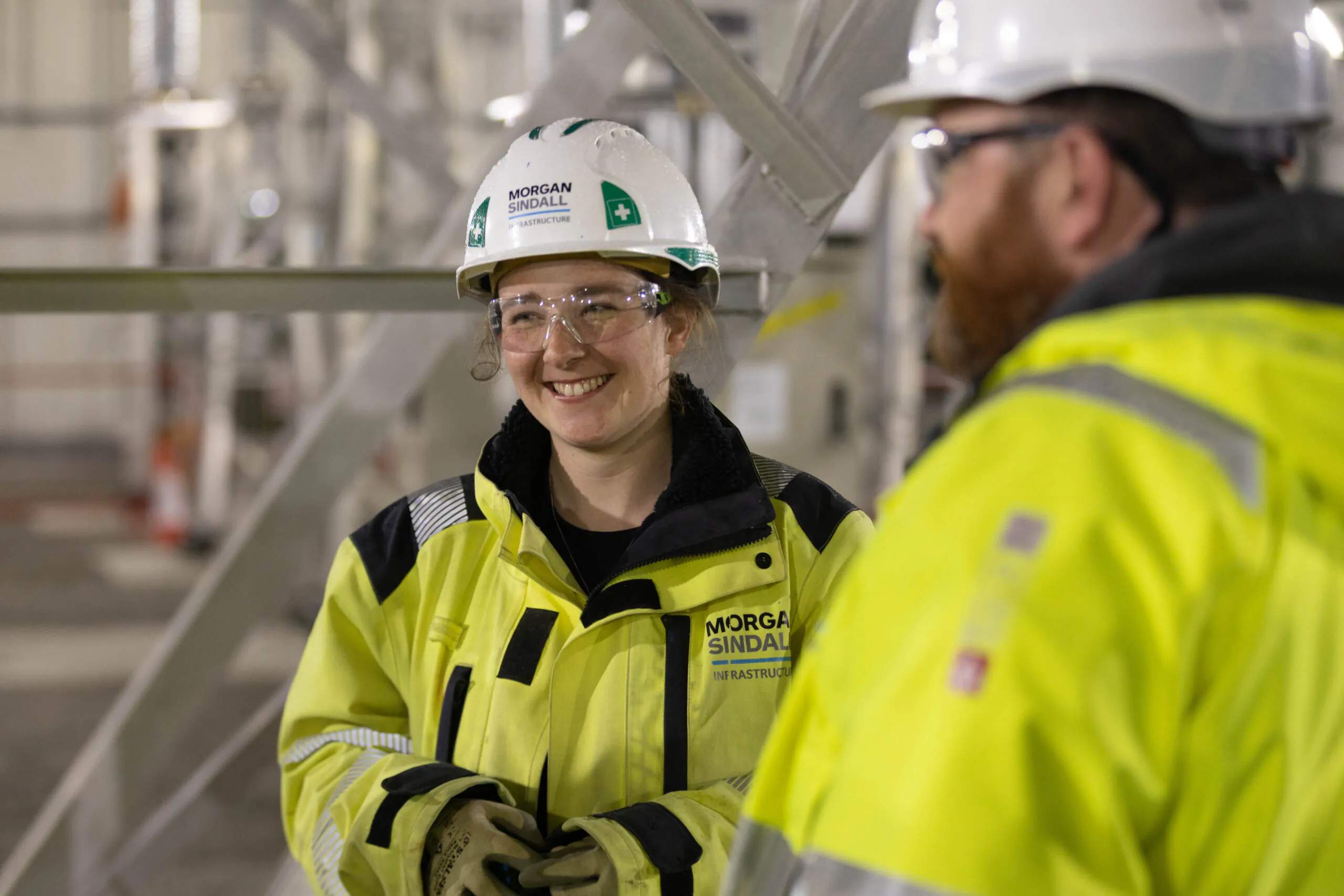 Two people in high-visibility jackets and white hard hats conversing indoors at an industrial or construction site.