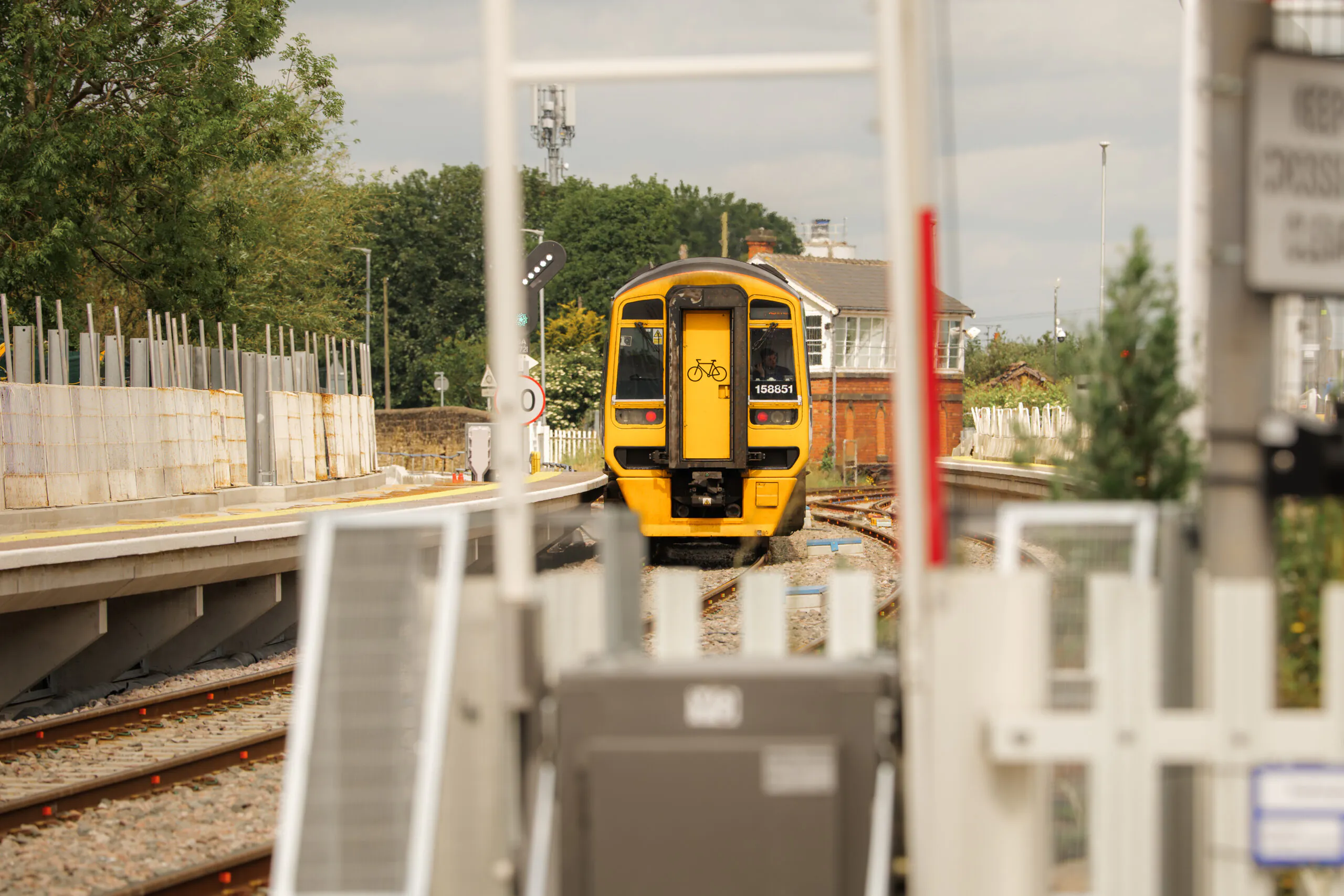 Yellow train approaching or departing an under-construction station platform during the day; safety barriers and signal lights visible.