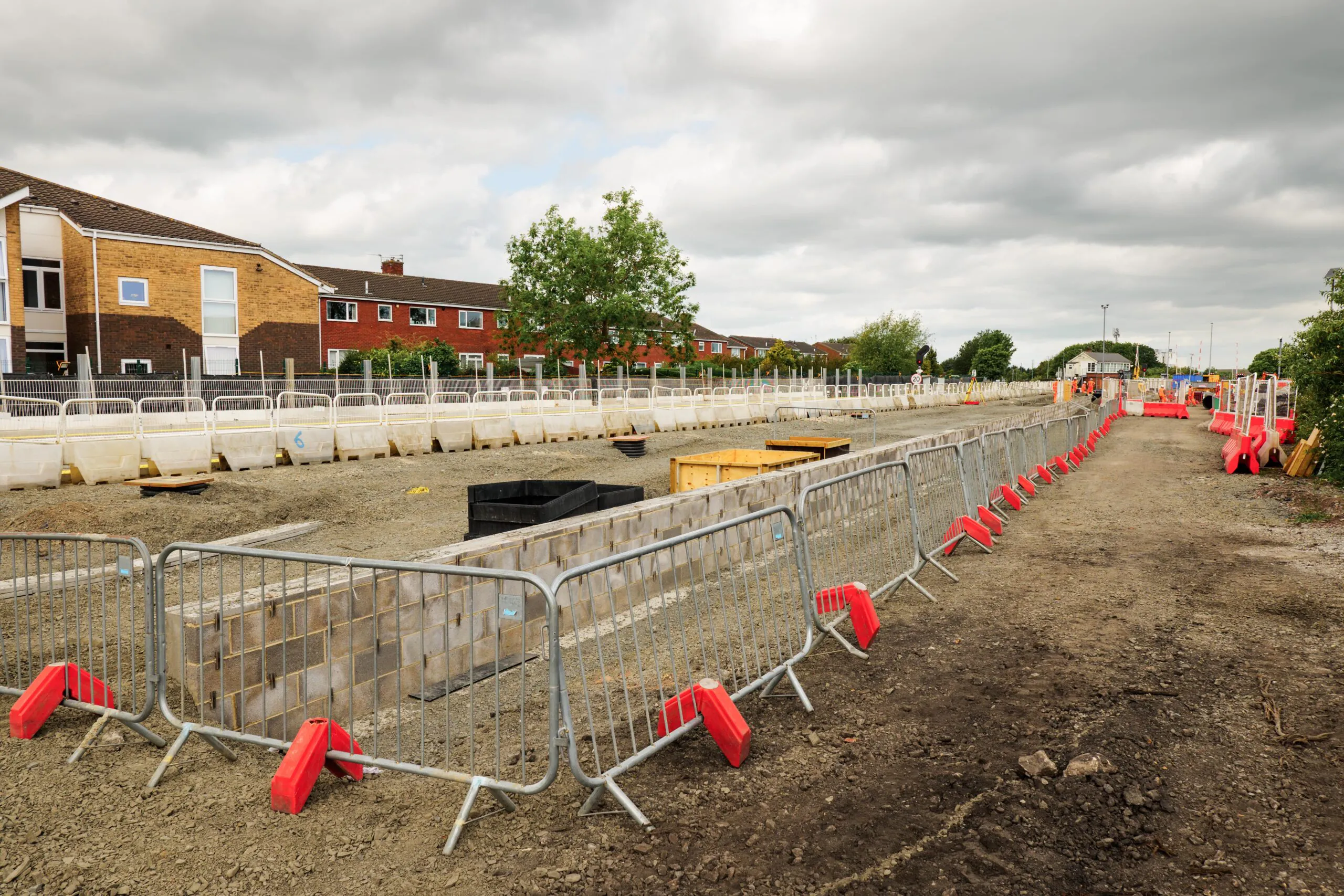 Daytime construction site with metal barriers and red plastic bases surrounding partially built brick structures; residential buildings visible in the background under a cloudy sky.