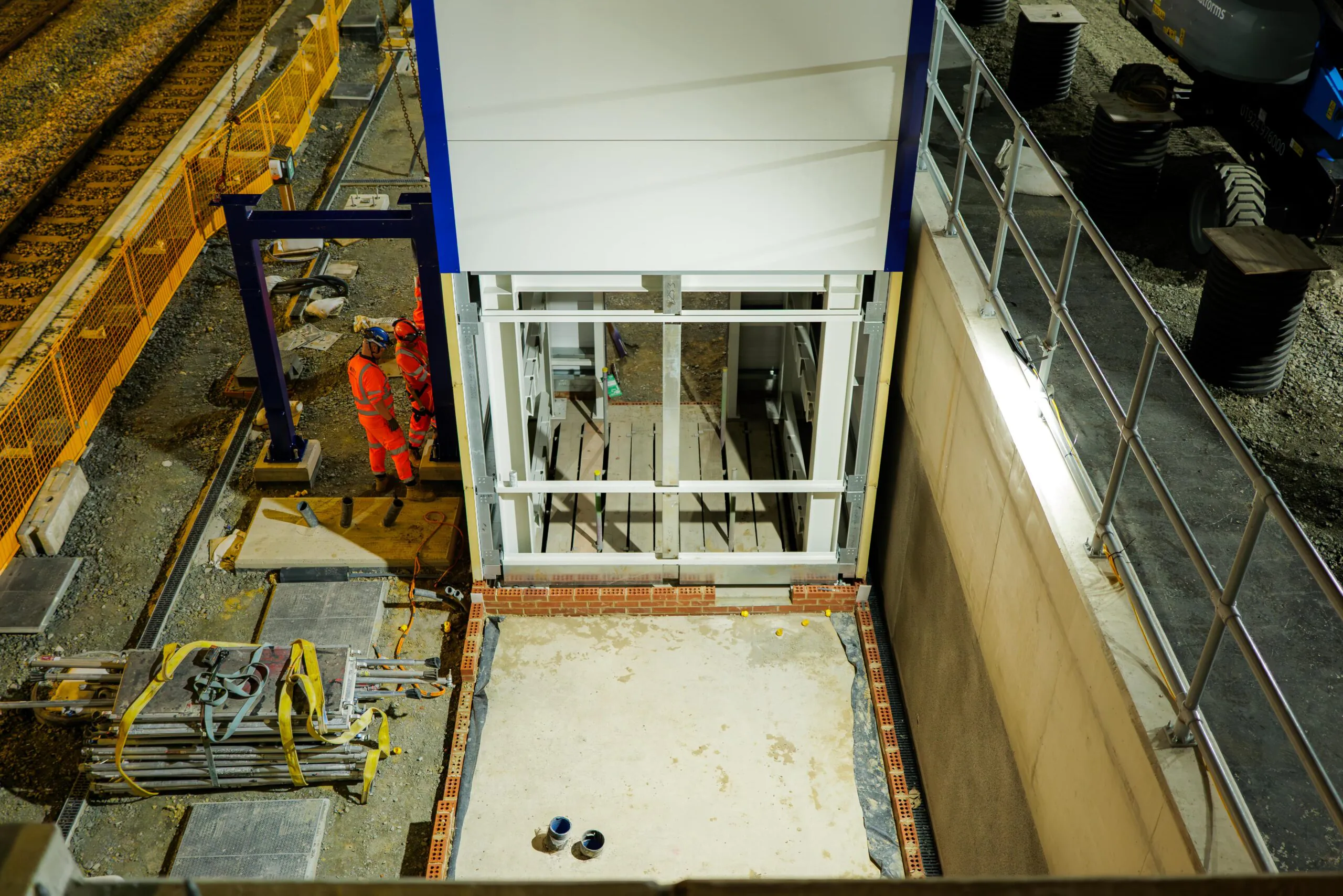 Overhead nighttime view of construction beside railway tracks; two workers stand inside a stairwell or elevator shaft structure illuminated by floodlights.