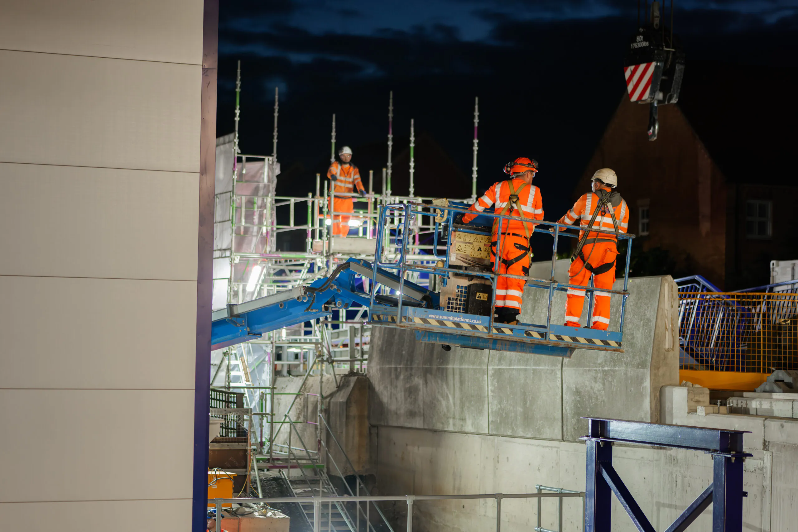 Nighttime construction scene with workers in high-visibility clothing operating machinery on scaffolding next to concrete structures.