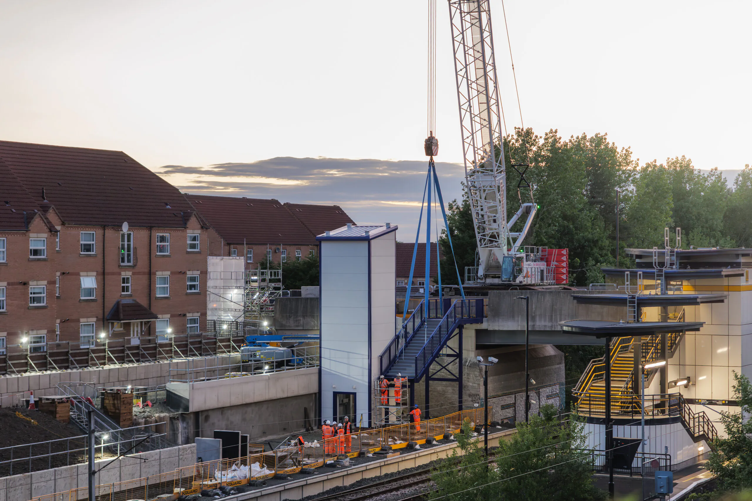 Wide-angle daytime view of railway station construction with cranes lifting materials and residential buildings nearby.