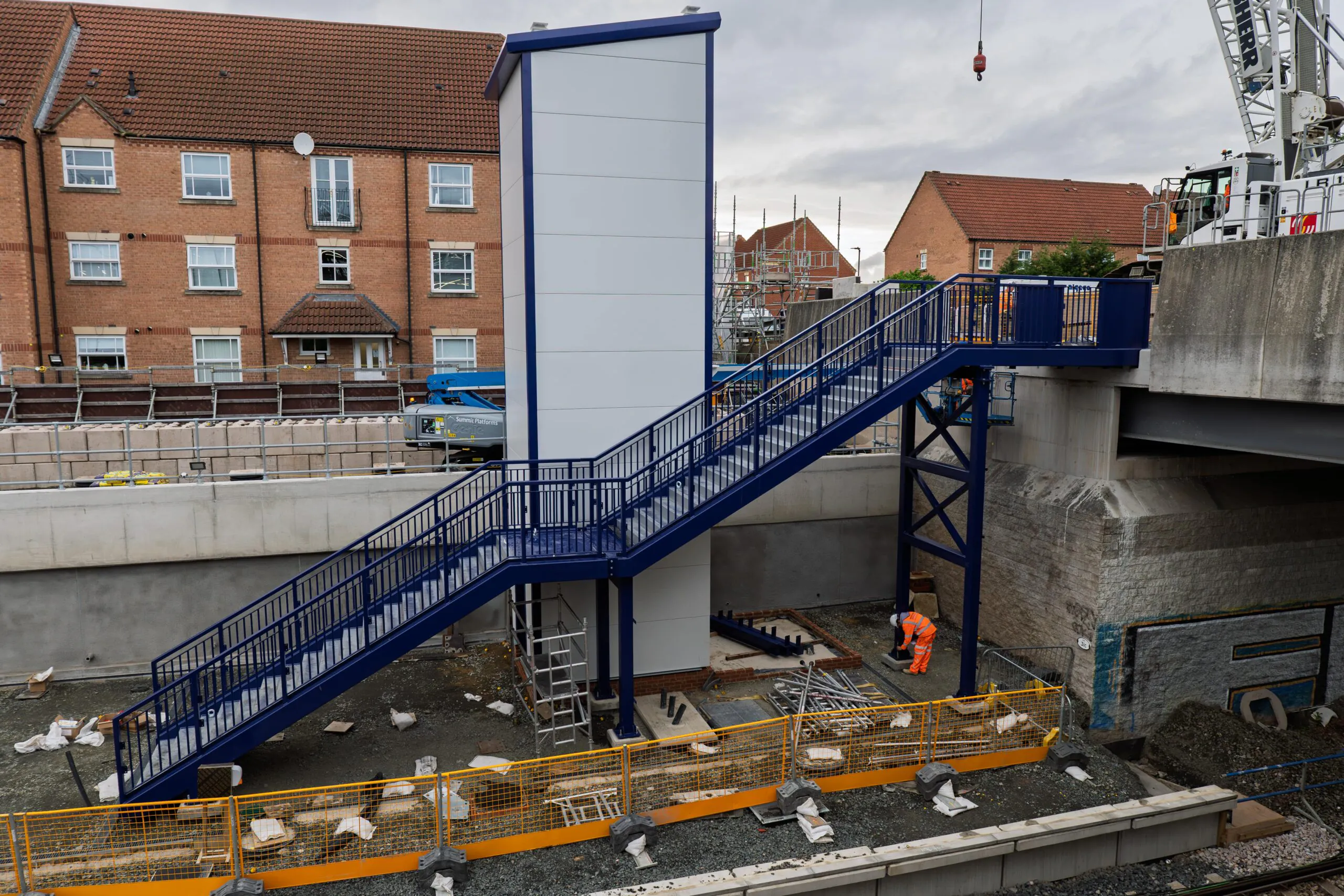 Daytime image of a nearly completed blue metal stairway structure at a railway construction site with workers present.
