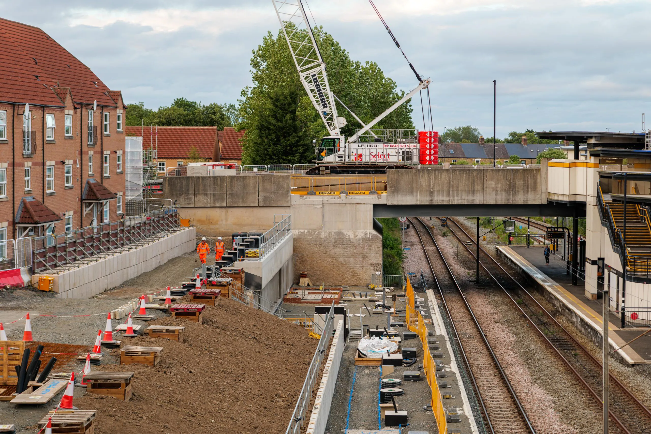 Daytime construction work beside railway tracks with cranes lifting materials onto platforms and workers below.