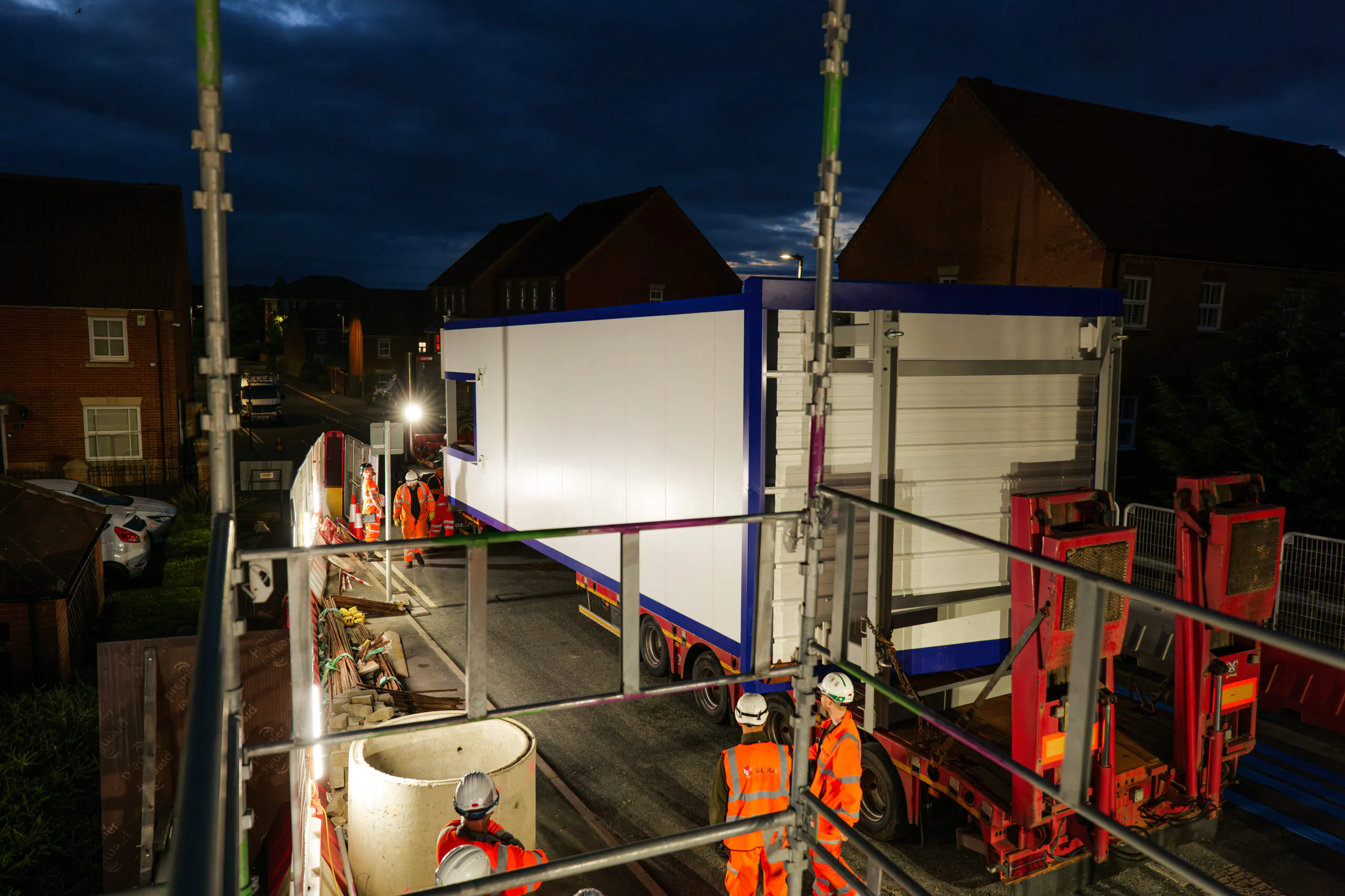 Nighttime construction scene featuring workers in high-visibility orange clothing and helmets operating around a large white modular building being transported on a flatbed truck. The site is illuminated by bright construction lights and surrounded by scaffolding, with residential houses visible in the background.