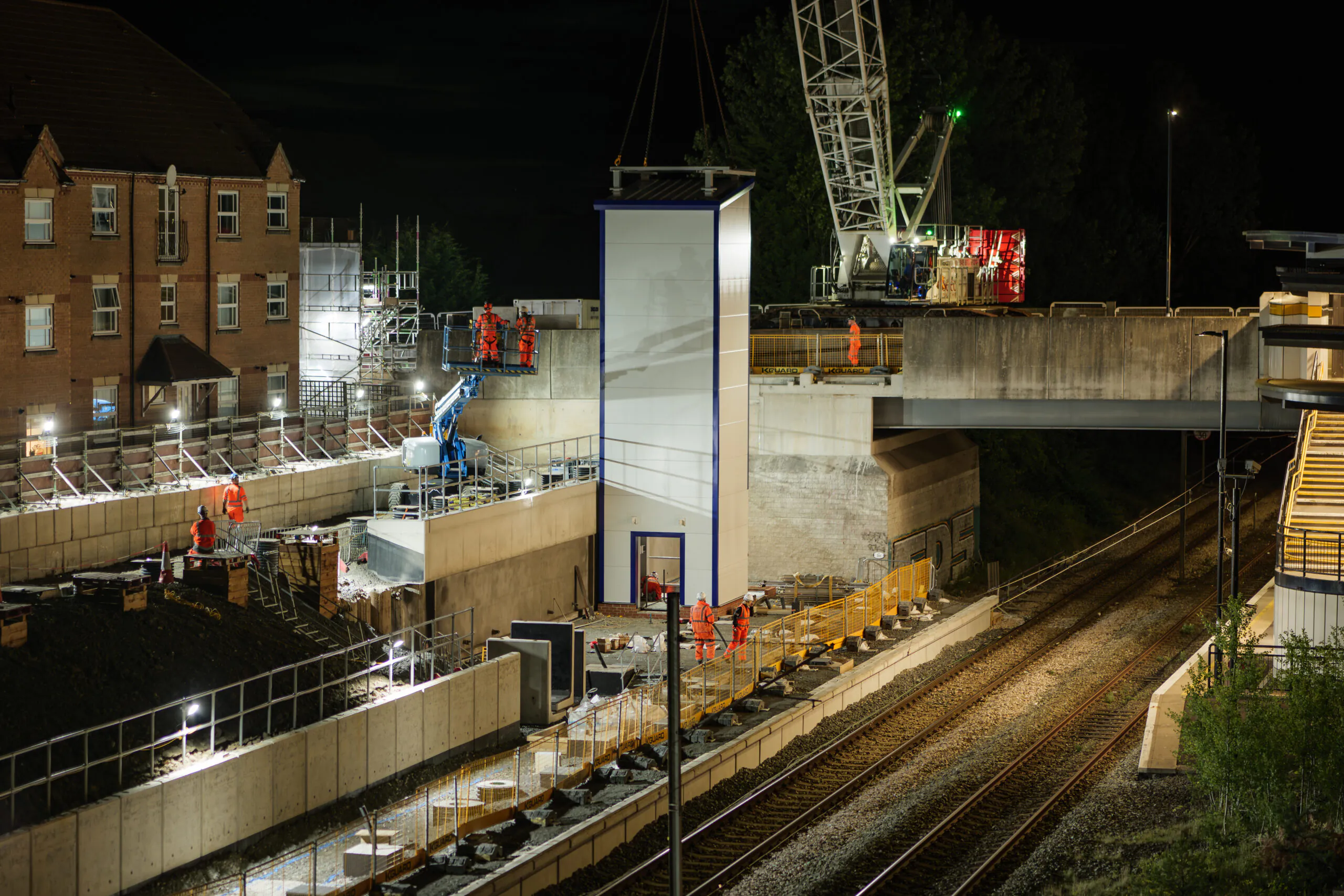 Nighttime overview of a railway station under construction with workers and equipment illuminated by bright lights.