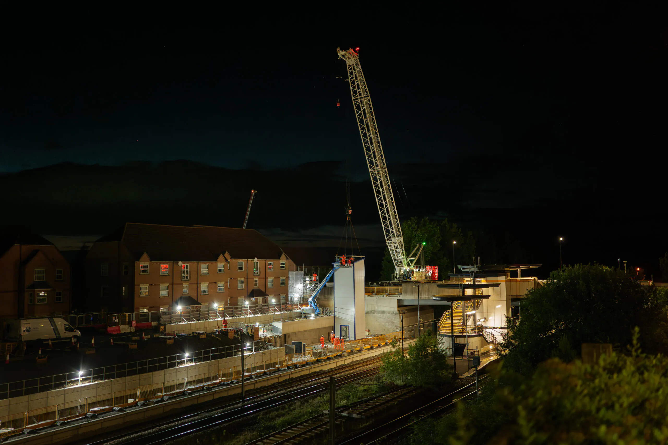 Elevated nighttime view of a construction site near railway tracks with a crane lifting components and workers on scaffolding; residential buildings in the background.