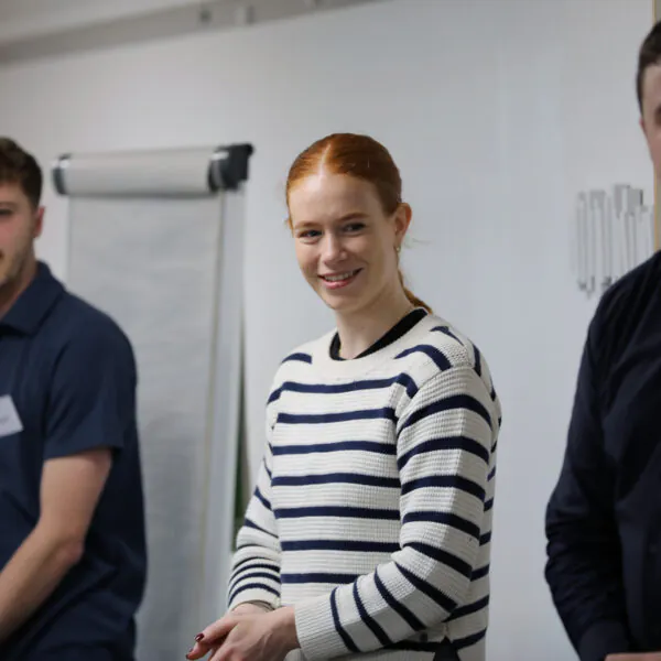 Participants stand during an induction or training session in a meeting room, with a flip chart and presentation space visible in the background.