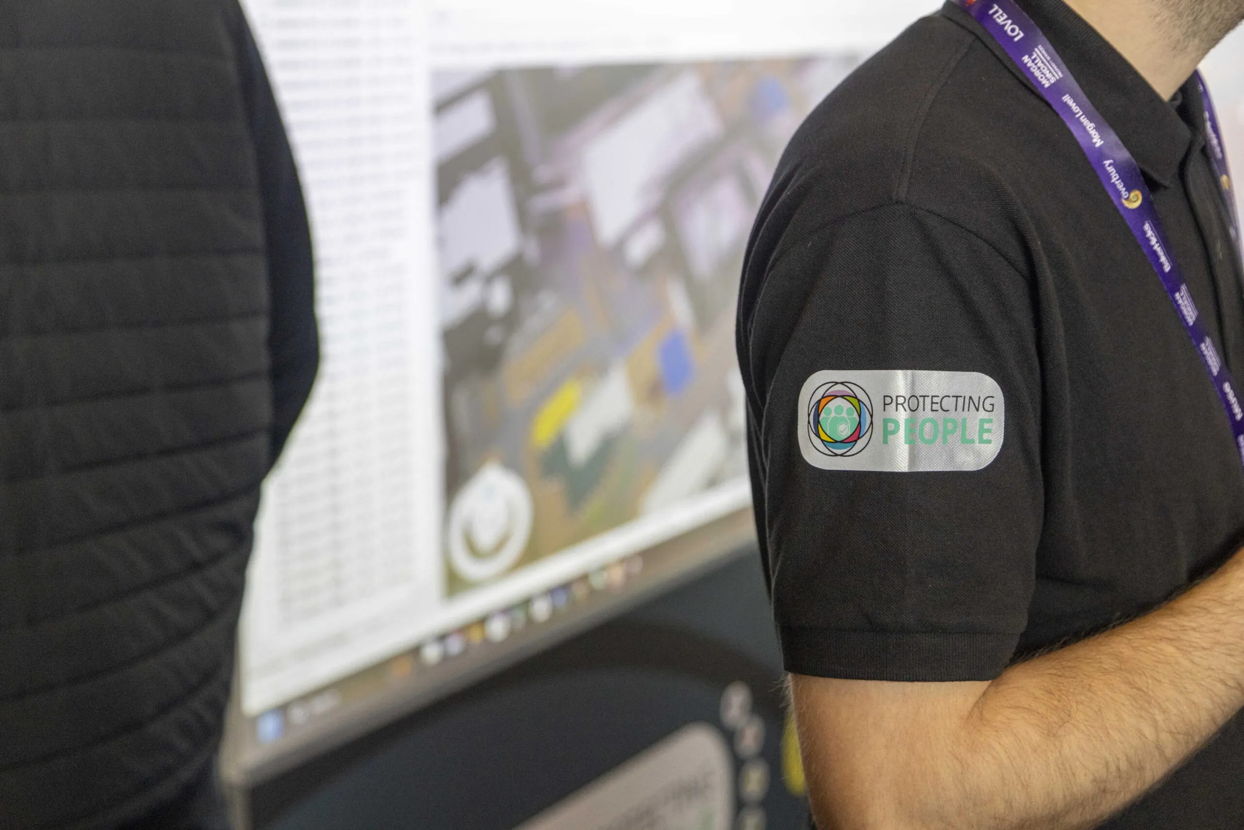 Close-up of two individuals wearing black shirts with a "Protecting People" emblem visible.