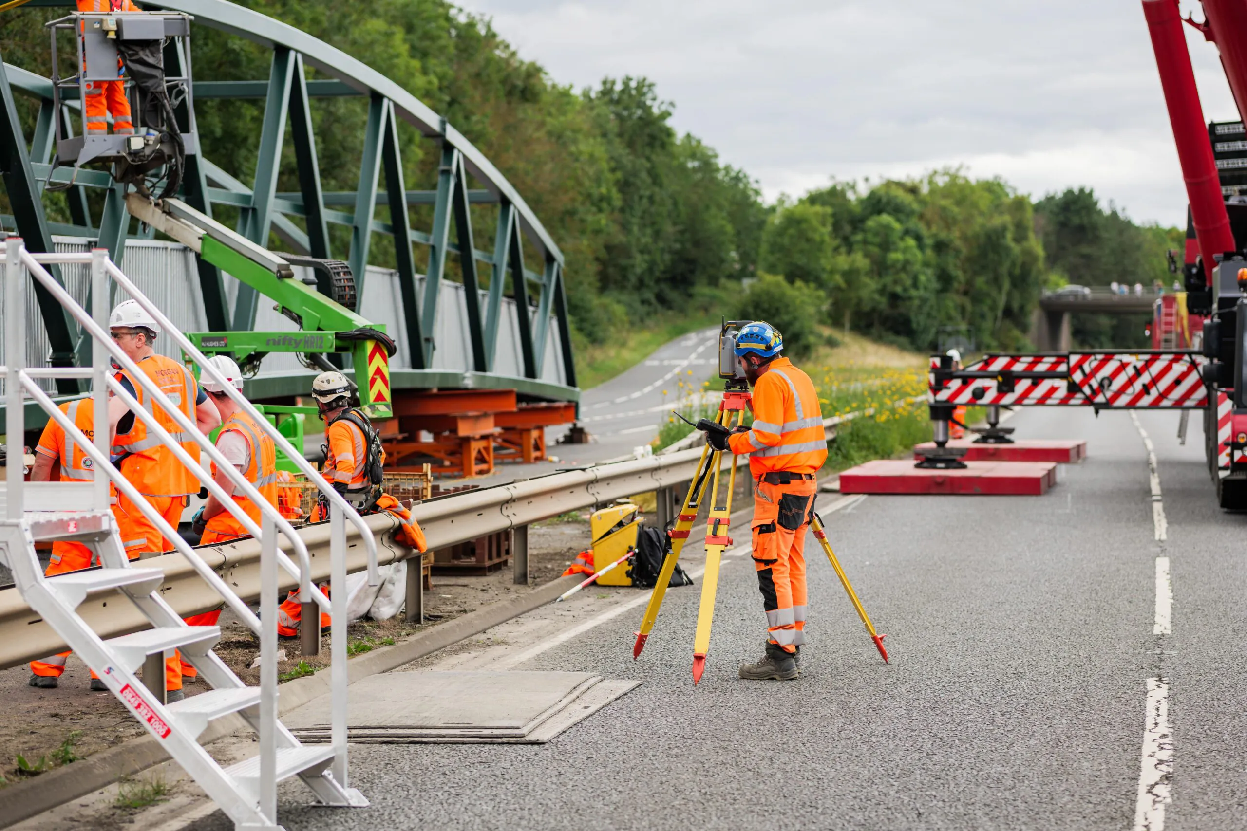 Construction workers in orange safety gear work near a green pedestrian bridge installation. One uses surveying equipment while others perform tasks around the site.
