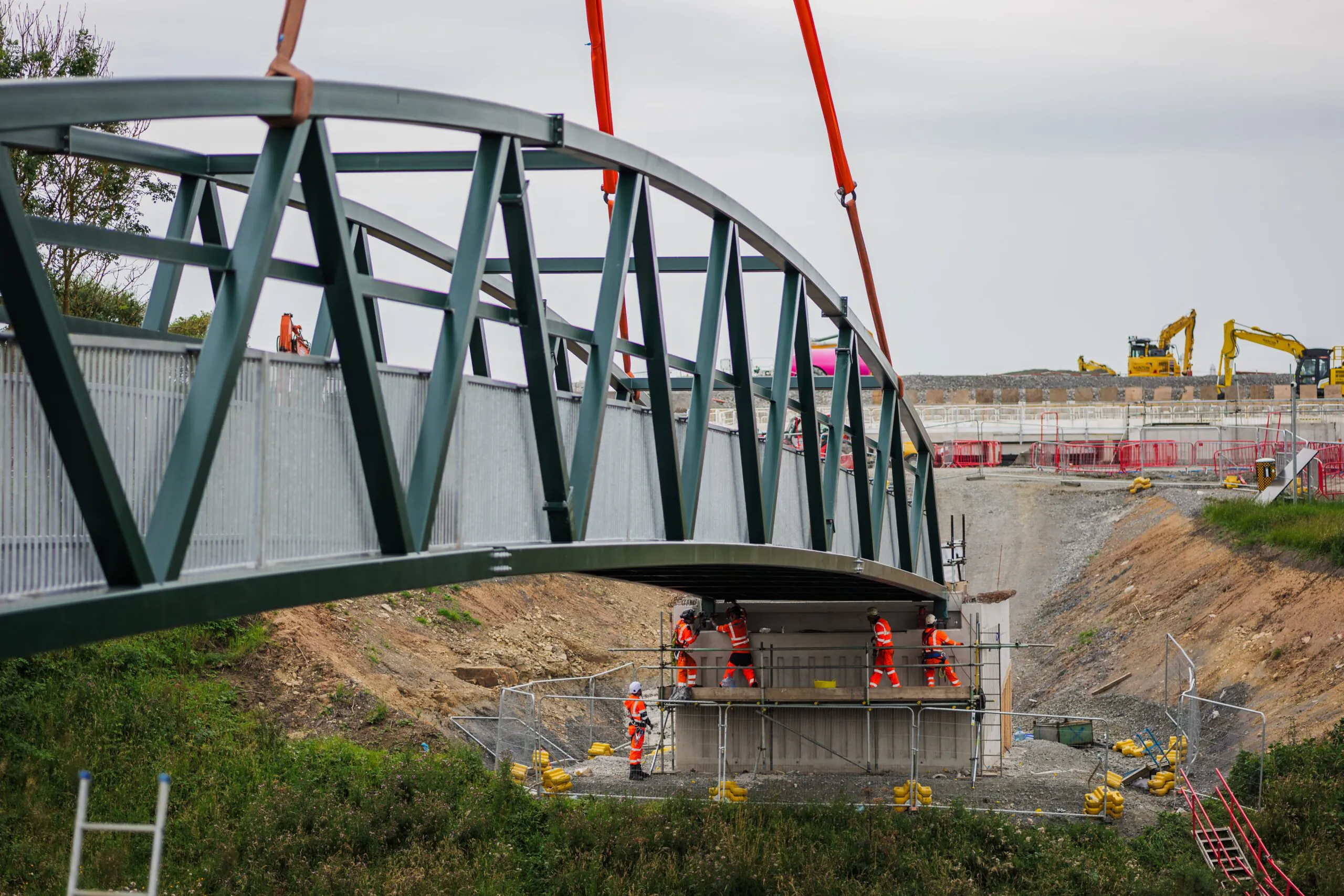 A large green pedestrian bridge is being installed over an excavation site. Workers in orange gear and cranes are actively involved in the construction process.