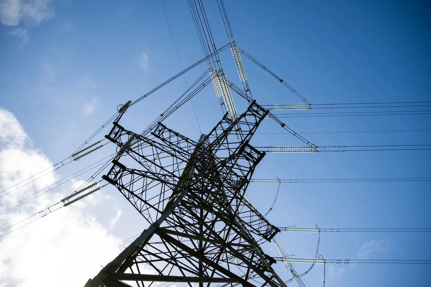 Upward view of a transmission tower with radiating power lines, under a mostly clear sky with some clouds.