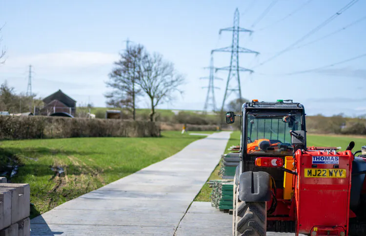 Red tractor on a paved path in a rural area, leading toward distant transmission towers, surrounded by green fields and trees.