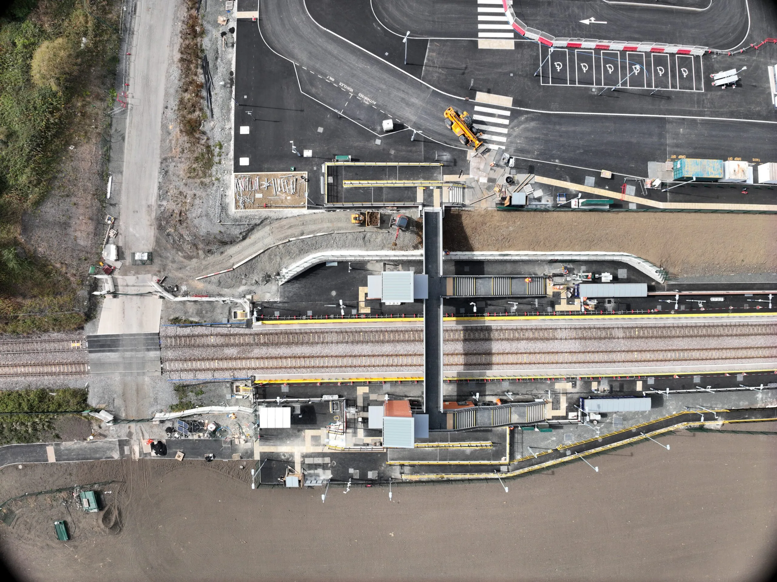 Top-down aerial view of a railway station construction site featuring multiple train tracks, platforms with shelters, pedestrian bridges, parking areas under development, and surrounding infrastructure.
