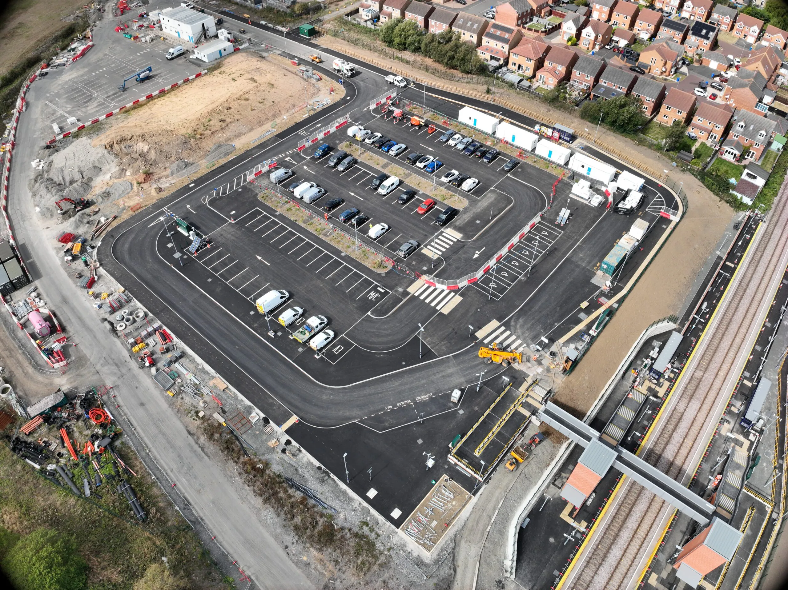 Aerial view of a large parking lot under construction, with construction materials, equipment, parked cars, temporary structures, and adjacent residential buildings.