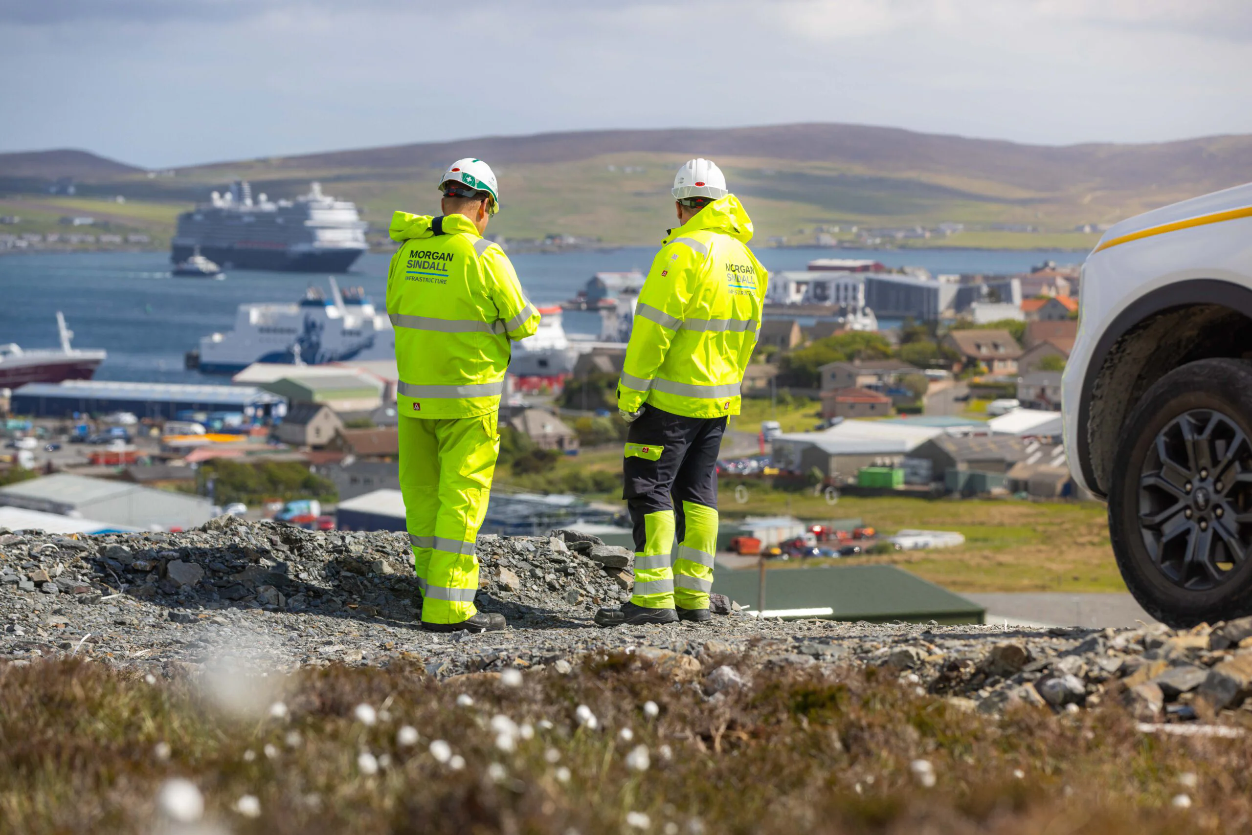 Two individuals in safety gear stand on a rocky hill overlooking a coastal town with docked ships; a Morgan Sindall-branded vehicle is partially visible.