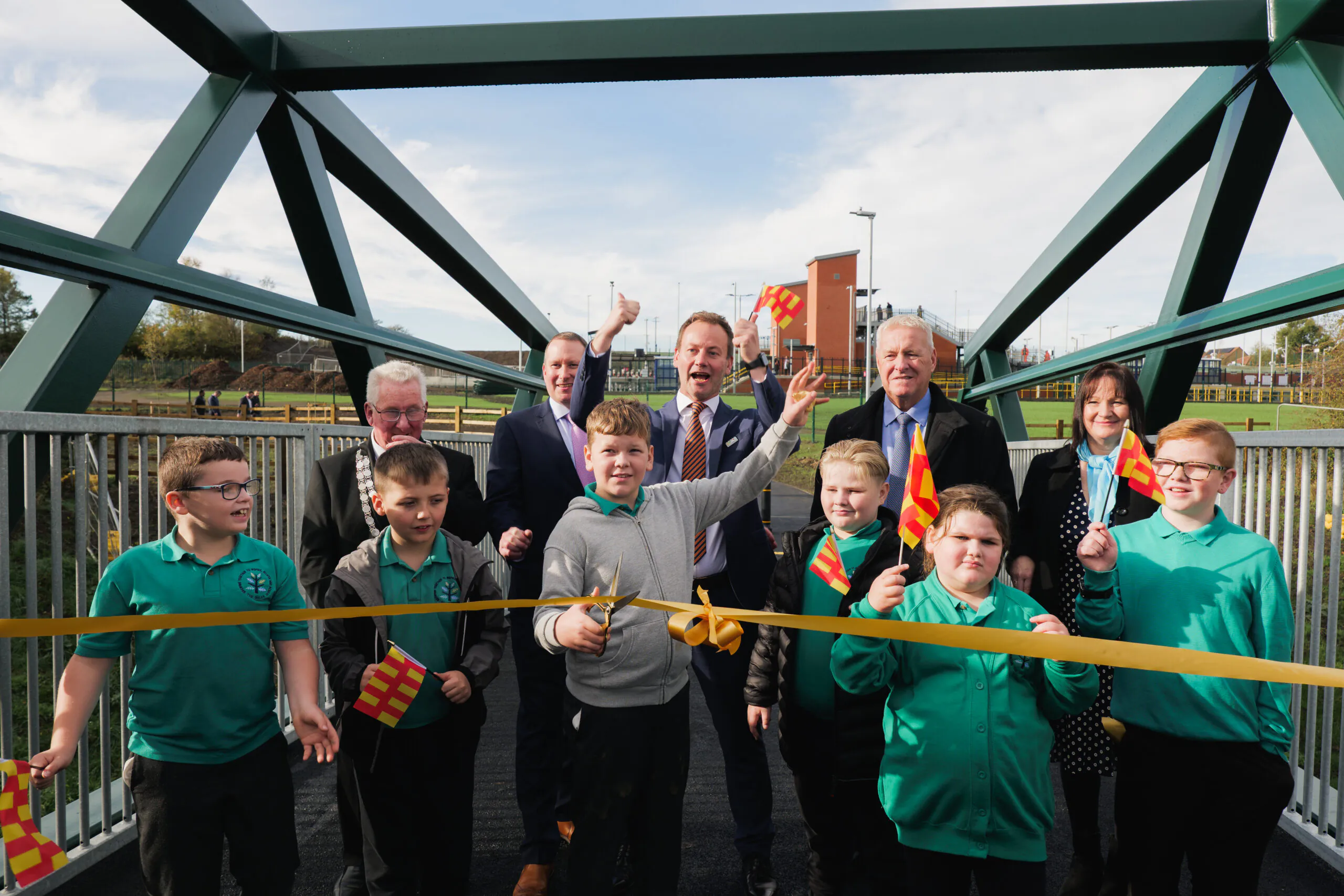 Children and adults gather on a pedestrian bridge holding small flags, participating in a ribbon-cutting ceremony with a yellow ribbon stretched across the bridge.