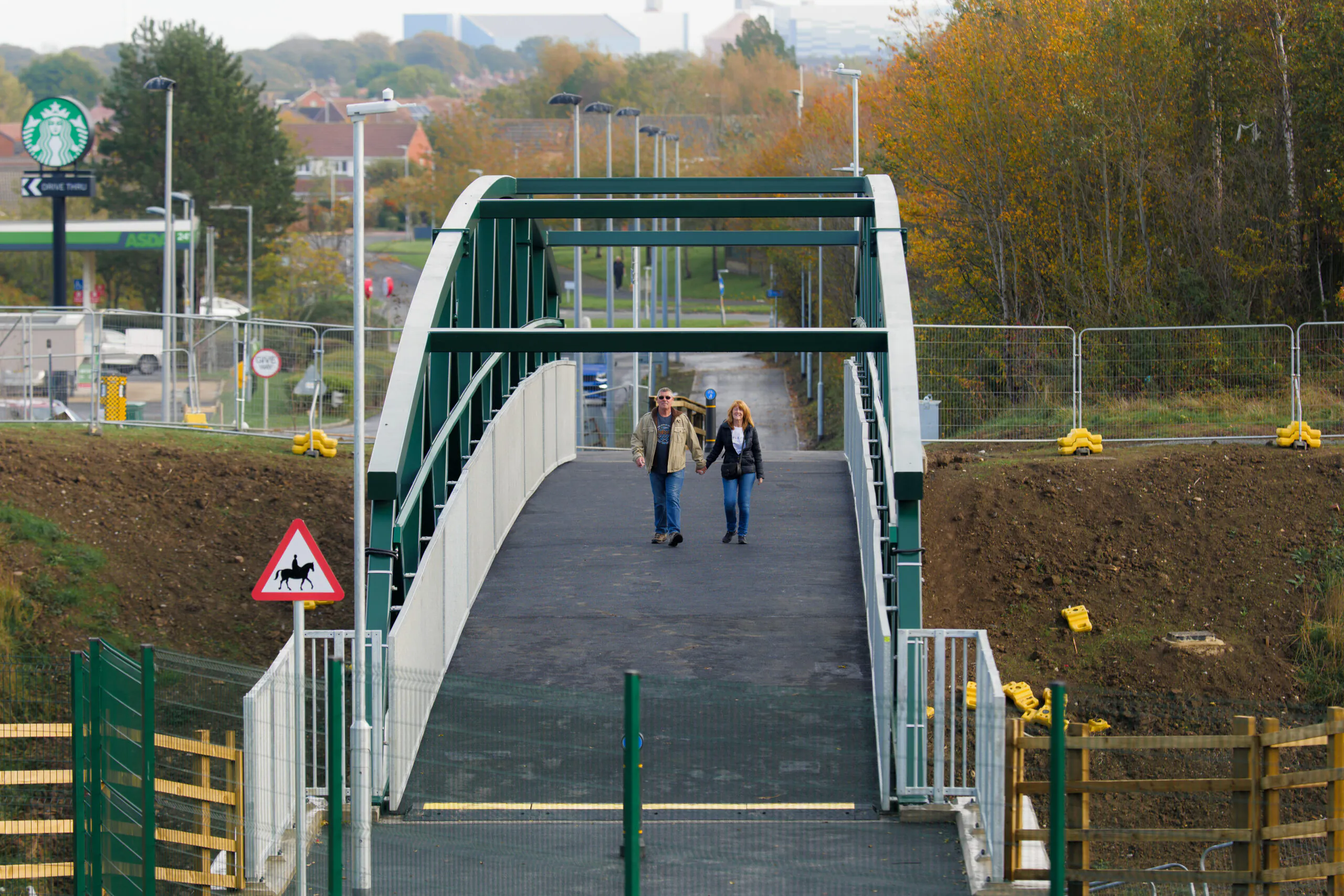 Two pedestrians walk across a green bridge with white railings above a road. A Starbucks sign and urban buildings are visible in the background.