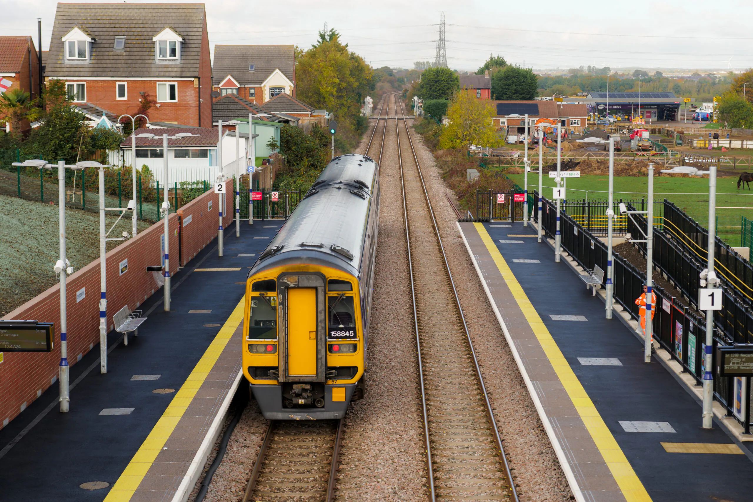 A train station with two platforms, where a yellow and grey train is stopped at platform 2. Platform 1 is empty, and the tracks stretch into the distance. Residential houses line the left side, while a green field with trees borders the right. The platforms feature benches, signs, and lights, with buildings and a power line tower visible in the cloudy background.