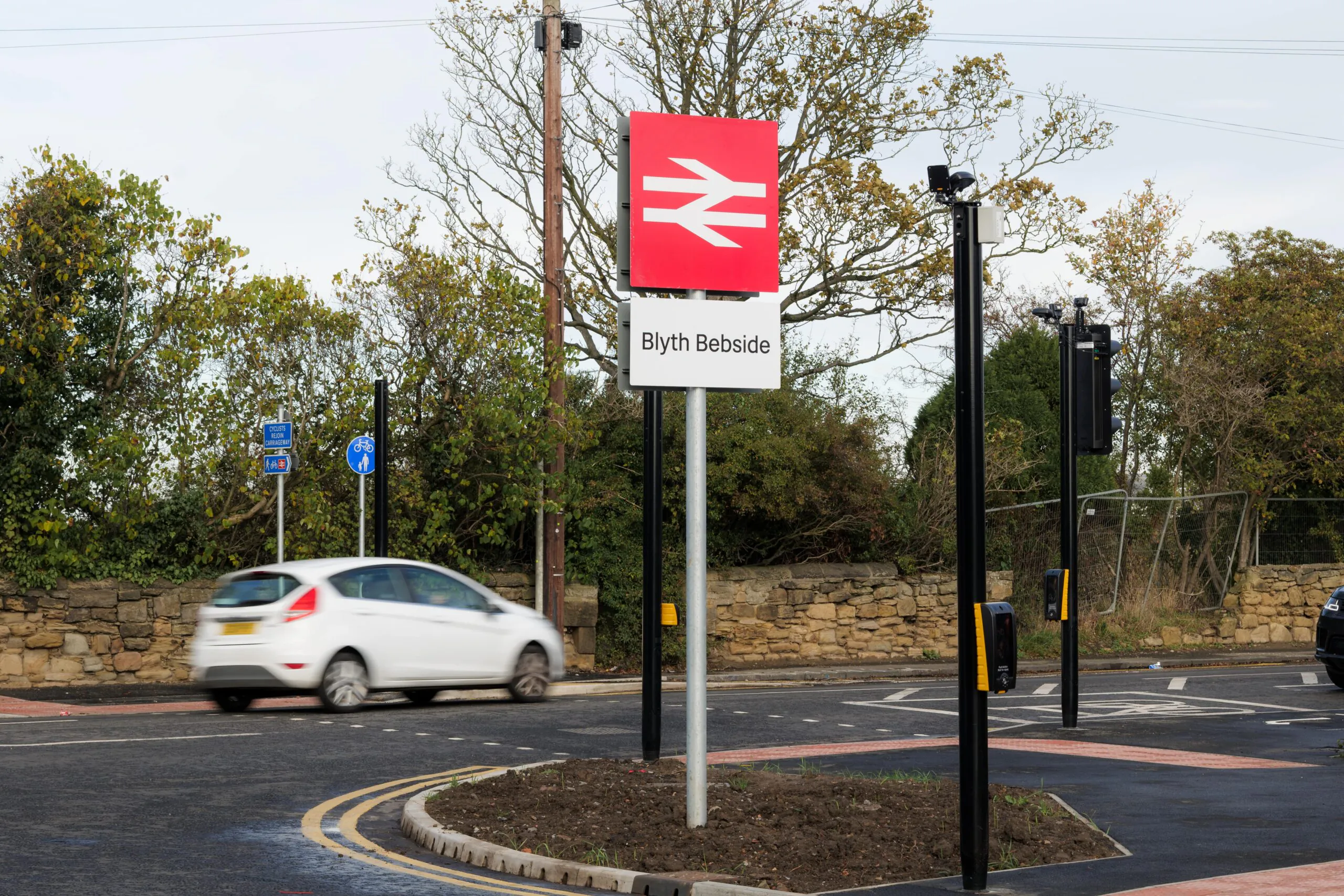 A roundabout featuring a road sign for "Blyth Bebside" with the British Rail logo. A white car navigates the roundabout, surrounded by trees and a stone wall.