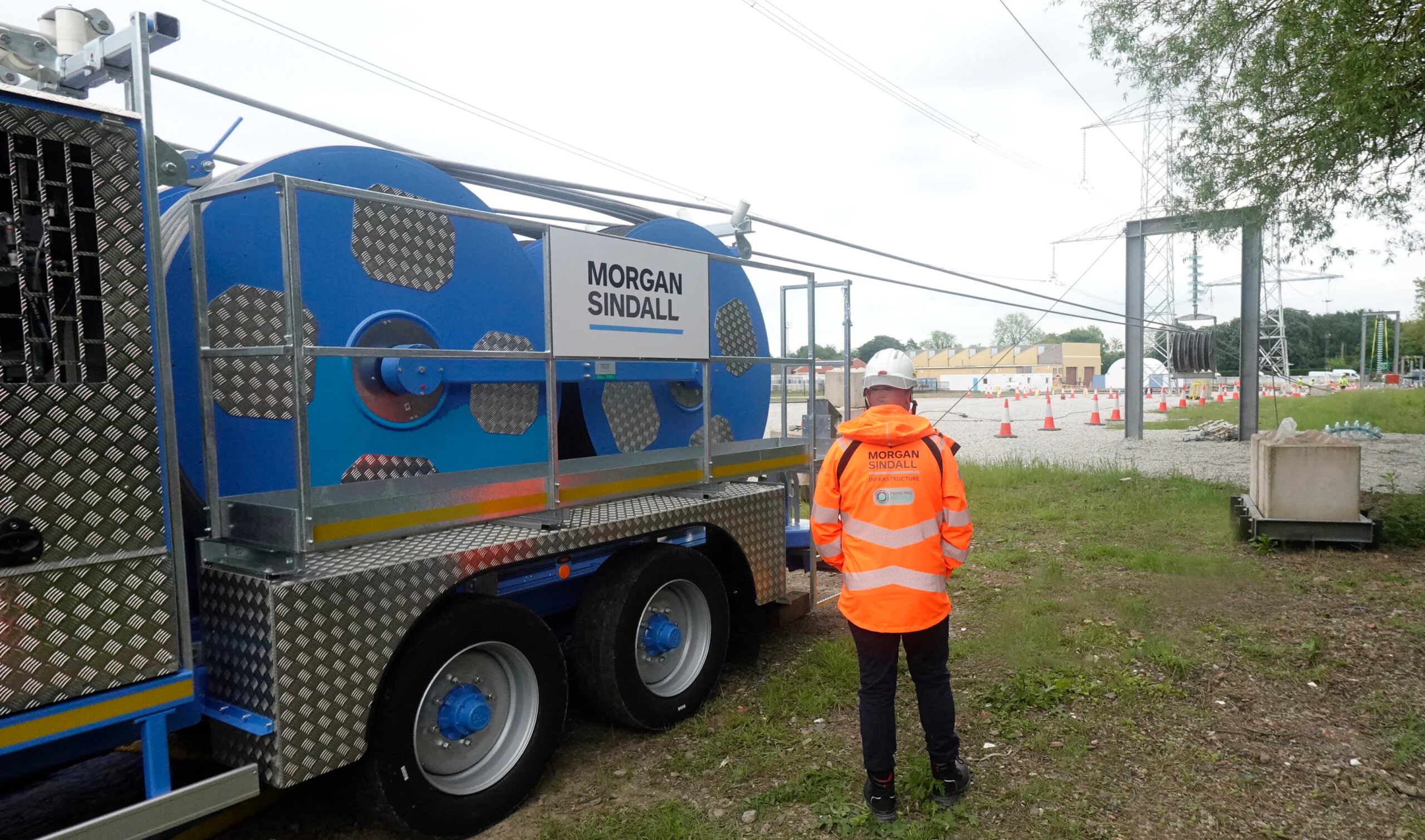 Worker in high-visibility gear standing beside large cable reels mounted on a trailer near power lines and traffic cones at an infrastructure site.