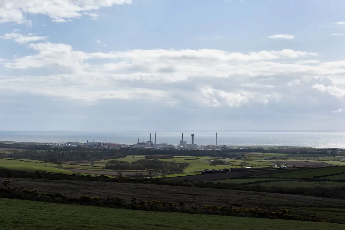 Panoramic view of a coastal industrial area with tall chimneys and buildings near the shoreline, surrounded by green fields and rolling hills in the foreground and the sea in the background under a partly cloudy sky.