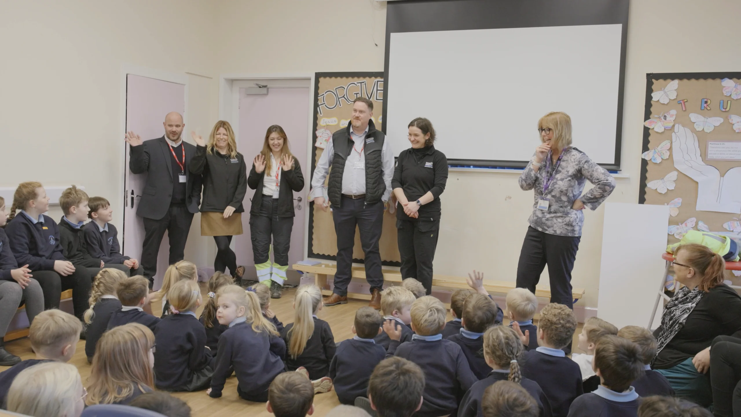 Adults stand at the front of a classroom introducing themselves to a group of seated schoolchildren in uniform. Several adults wear branded work jackets and lanyards, with one raising both hands in greeting. A teacher stands to the side near a whiteboard and wall displays featuring student work and a board titled “FORGIVE.” The children sit on the floor facing the speakers, with some raising their hands.